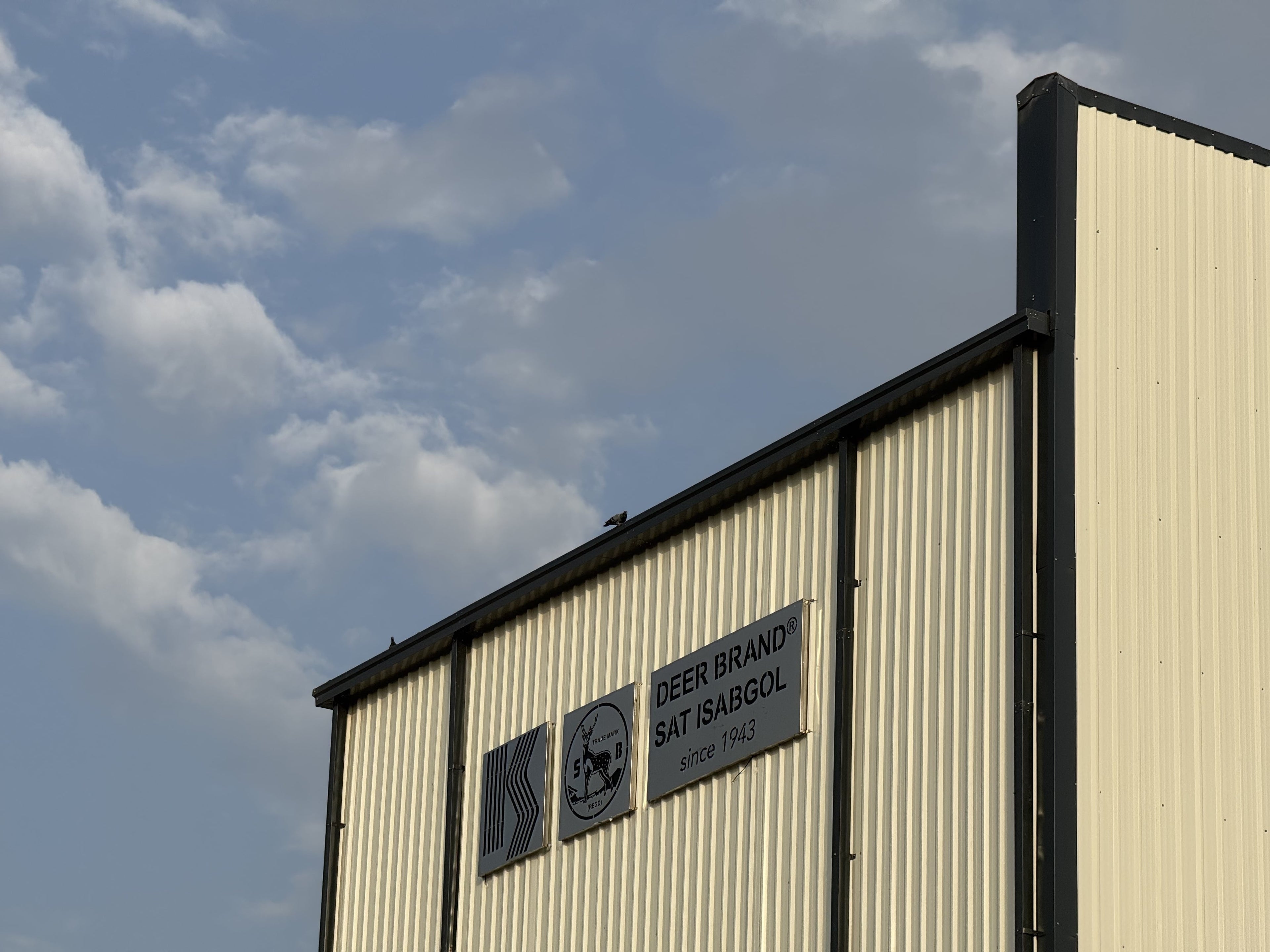 A close-up of a corrugated metal building exterior with a cream-colored panel wall, black trim, and a sign reading 'DEER BRAND SAT ISABOL' against a blue sky with clouds.